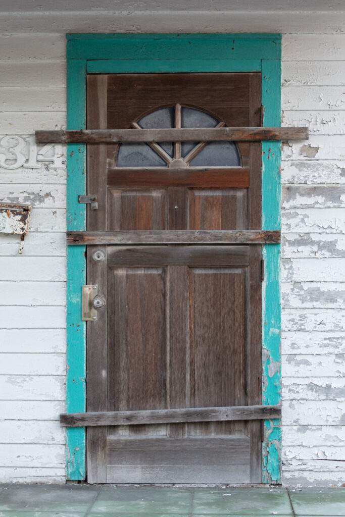 “Boarded wooden door on a weathered residential structure with peeling paint and turquoise trim.”